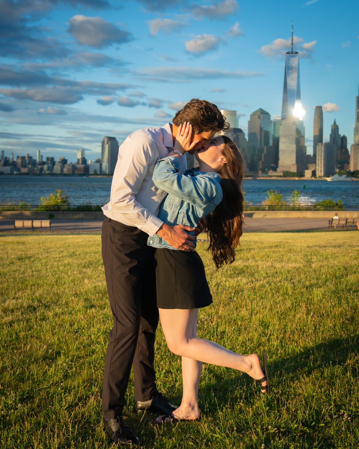 A couple kissing on a riverside lawn at golden hour, the Lower Manhattan skyline rising behind them.