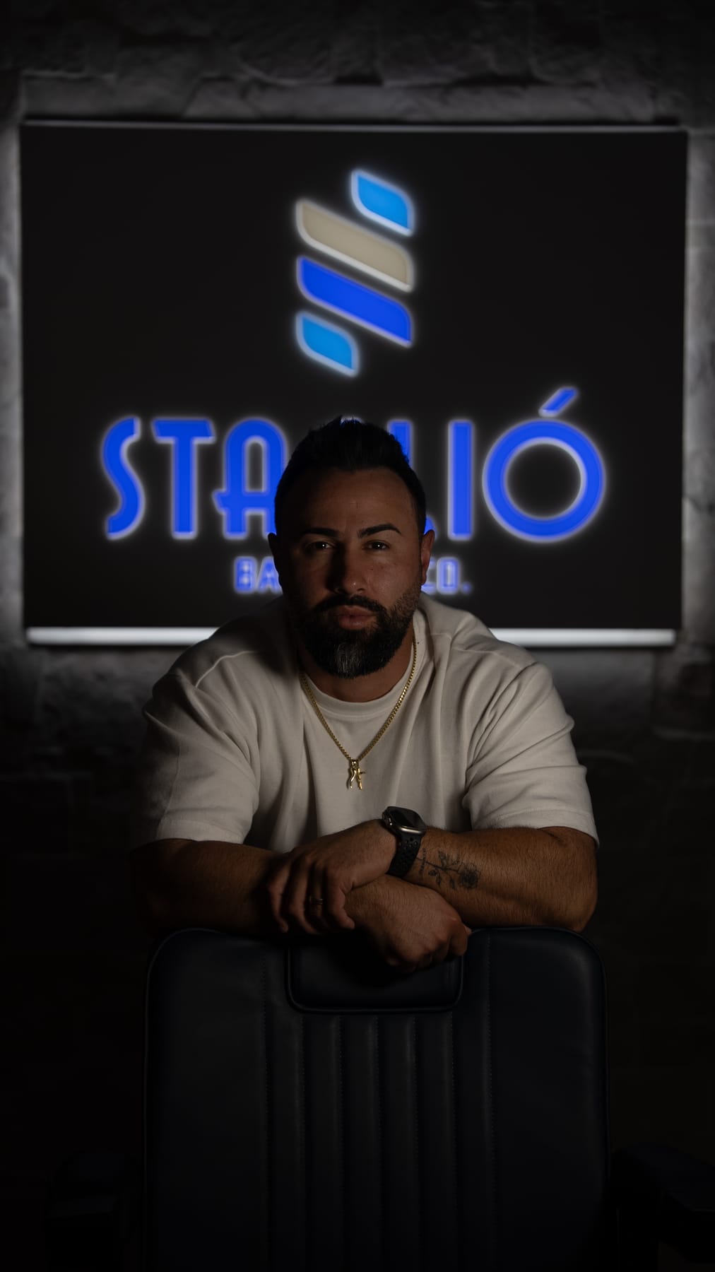 A low-lit portrait of a bearded man leaning on a barber chair in front of an illuminated Stagliò sign.