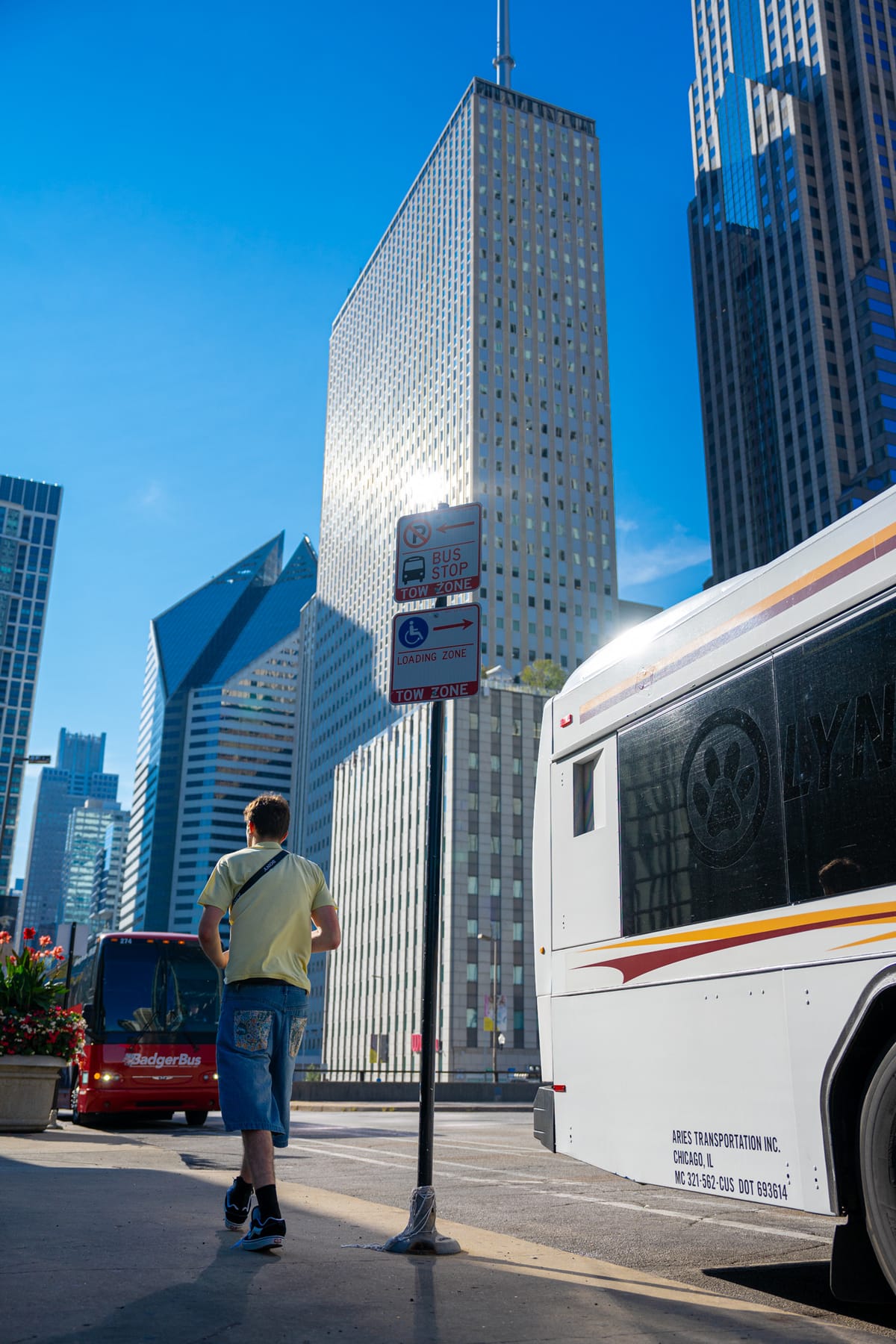 A man walking toward a downtown Chicago bus stop, coach buses framing him and skyscrapers overhead.