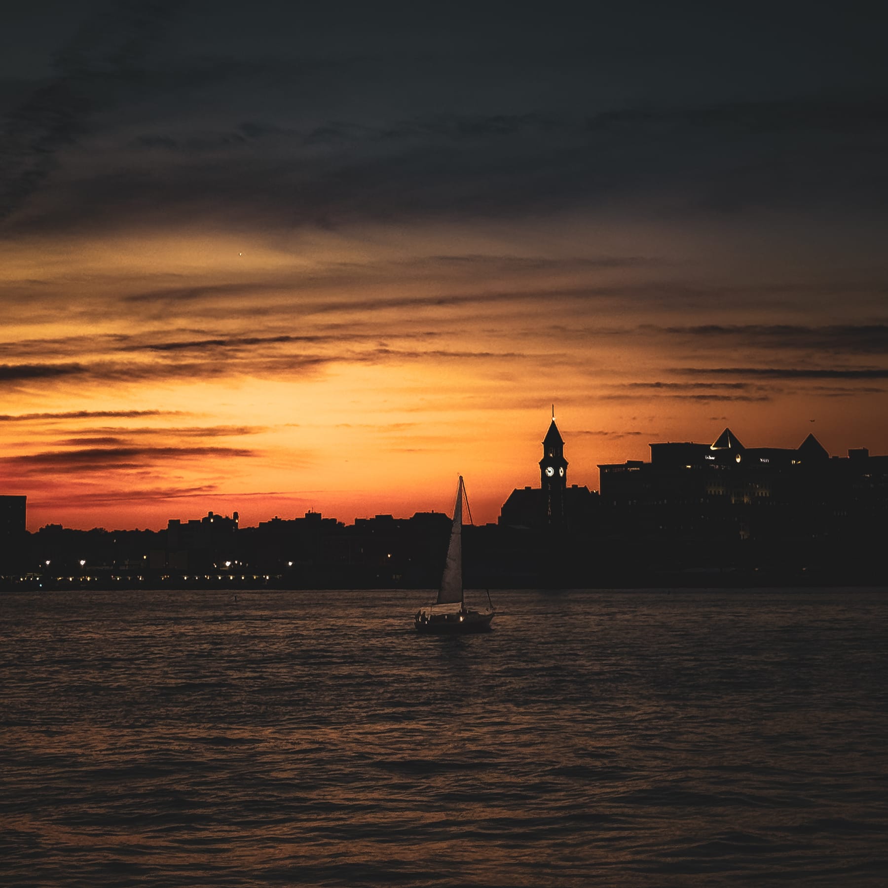 A lone sailboat crossing the Hudson at dusk, the Hoboken clock tower silhouetted against a deep orange sky.