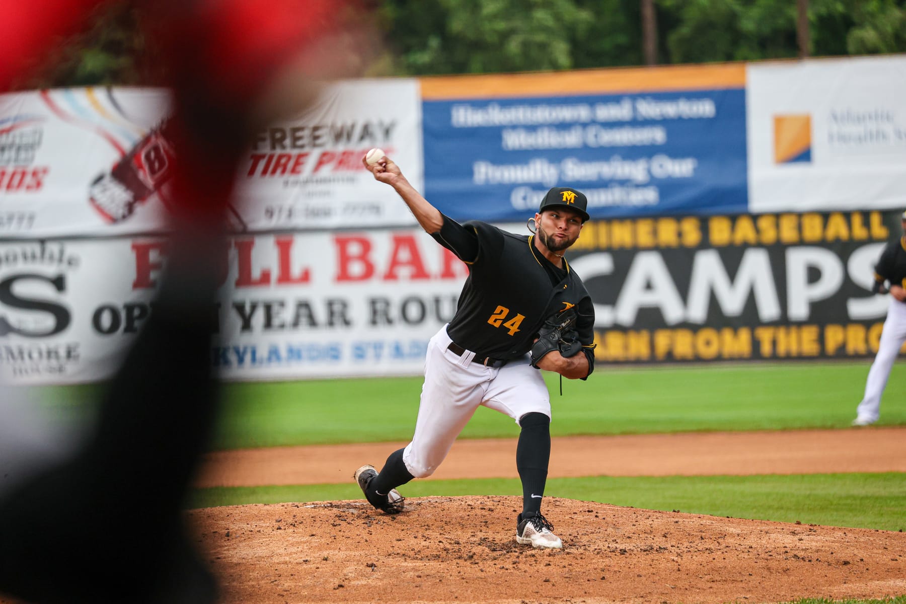 A baseball pitcher in a black and gold uniform at full extension mid-delivery, ball just leaving his hand.