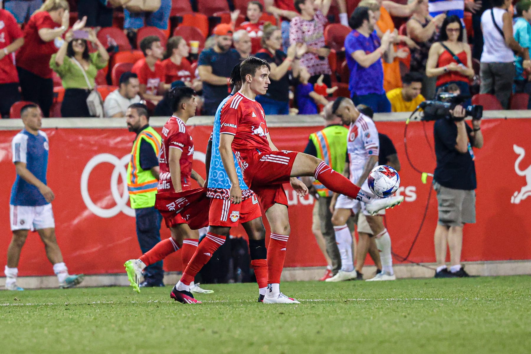 A New York Red Bulls player juggling the ball on the pitch after a match while the crowd applauds behind him.